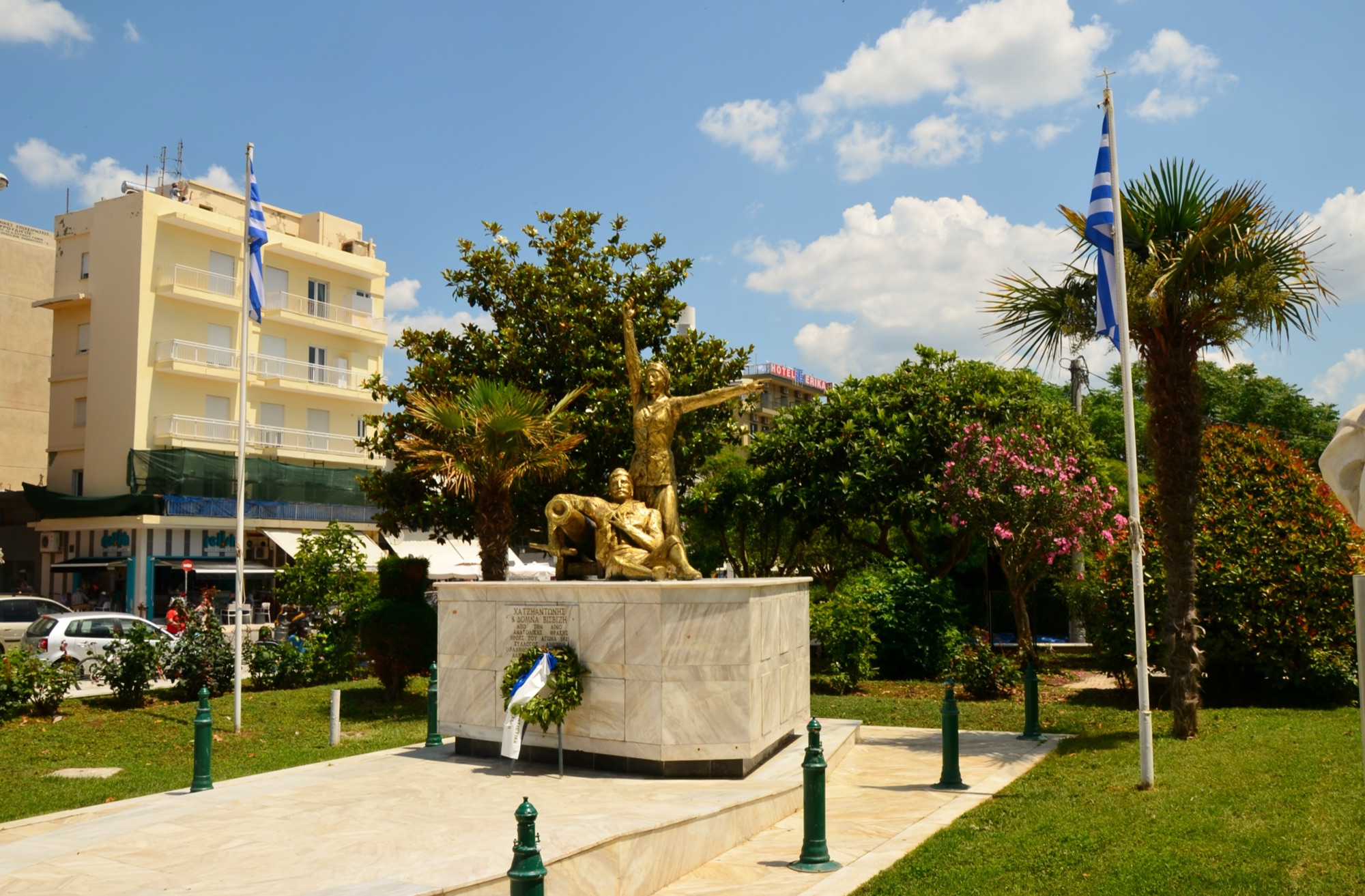 A war memorial statue framed by palm trees and Greek flags in central Alexandroupolis