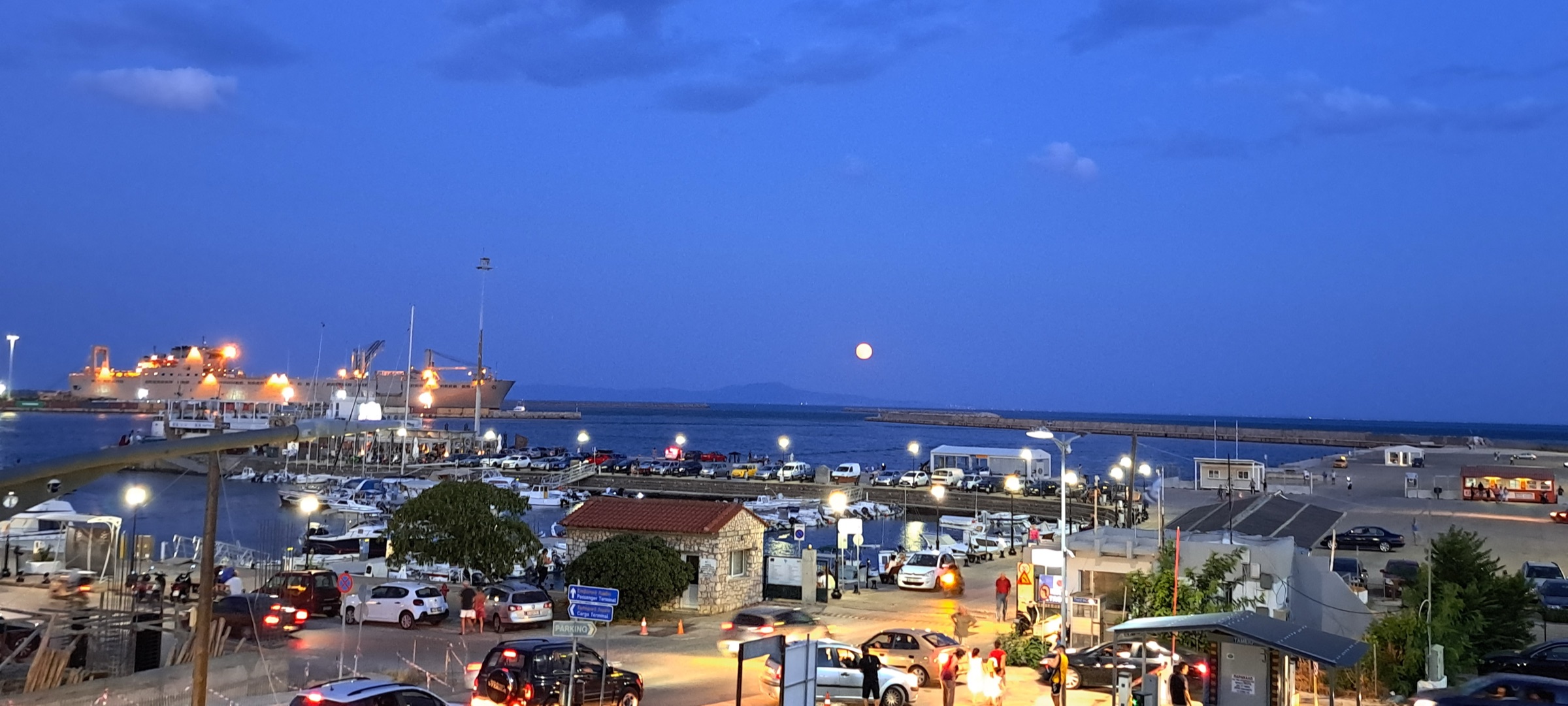The Alexandroupolis harbour at blue hour, with a lit ferry, string lights along the pier, and the moon rising over the sea